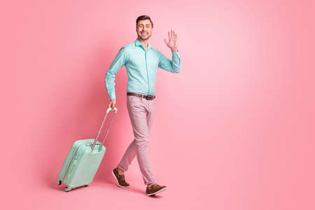 Full size profile side photo of brunette man go wave hold luggage wear teal shirt pink pants isolated on pastel color backgroundの写真素材