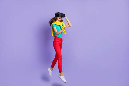 Full length body size view of skinny cheerful girl jumping wearing vr helmet playing game isolated over violet pastel color backgroundの写真素材