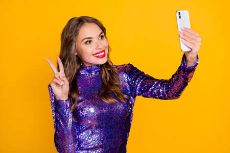 Photo portrait of smiling woman taking selfie showing v-sign two fingers in festive dress isolated vibrant yellow color backgroundの写真素材