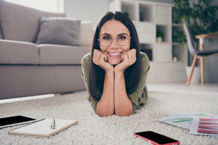 Portrait of charming cheerful person lying on floor hands on cheeks look camera toothy smile working from home indoorsの写真素材