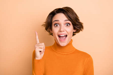 Photo of funny clever young lady wear orange turtleneck having idea pointing finger isolated beige...