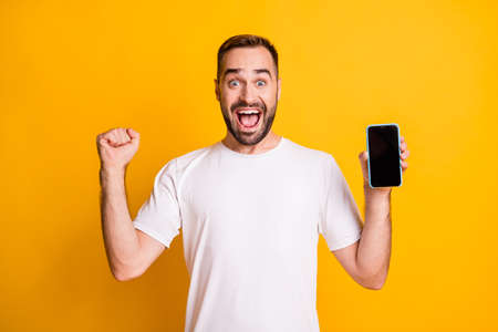 Portrait of attractive cheerful guy holding in hand device celebrating sale having fun isolated over bright yellow color backgroundの写真素材