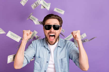 Photo of impressed lucky young guy dressed denim shirt dark glasses money flying rising fists isolated purple color backgroundの写真素材