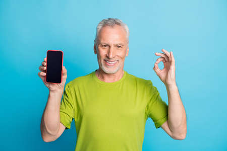 Portrait of attractive cheerful grey-haired man showing ok-sign holding in hand device isolated over bright blue color backgroundの写真素材