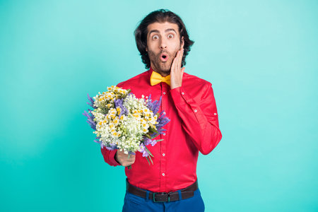 Photo of shocked young brown haired man hold hand cheek flowers present isolated on teal color backgroundの写真素材