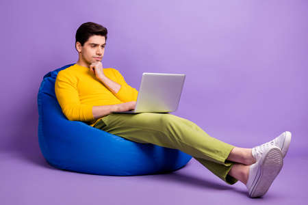 Full body profile side photo of young man serious think look laptop programmer sit chair bean isolated over violet color backgroundの写真素材