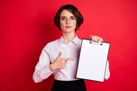 Photo of serious businesswoman in glasses point finger organizer attention announcement isolated on red color backgroundの写真素材