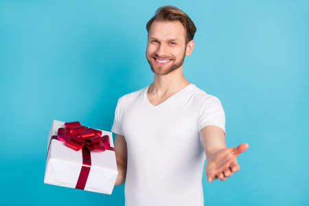 Photo of friendly candid man hold giftbox raise hand palm invite you toothy smile isolated on blue...