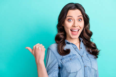 Photo of charming excited young lady dressed denim shirt smiling pointing thumb up empty space...