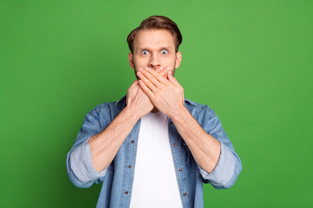 Photo of young shocked amazed surprised man cover mouth with hands oops guilty isolated on green...
