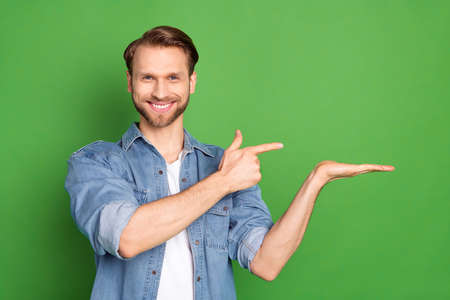 Photo portrait of young guy pointing blank space holding on palm surprised smiling isolated on vibrant green color backgroundの写真素材
