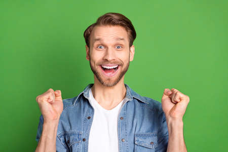 Photo portrait of young guy smiling in denim shirt gesturing like winner isolated on bright green color backgroundの写真素材