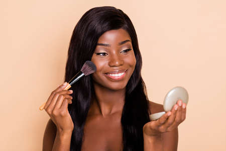Portrait of attractive cheerful shine girl applying powder on cheek preparing isolated over beige...