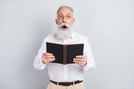 Photo of amazed shocked old man hold book surprised plot wear glasses isolated on grey color...