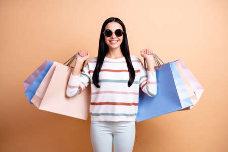 Photo of young beautiful cheerful smiling positive girl in sunglasses hold shopping bags isolated on beige color backgroundの写真素材
