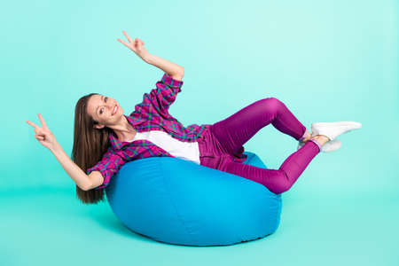Portrait of attractive cheerful funky girl lying in chair showing v-sign having fun isolated over bright teal color backgroundの写真素材