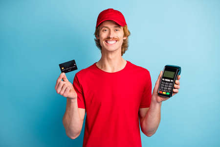 Photo of positive deliveryman hold banking card pos terminal smile good mood isolated on blue color...