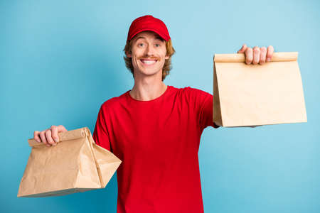 Portrait of satisfied person arms holding food orders toothy smile wear hat isolated on blue color backgroundの写真素材