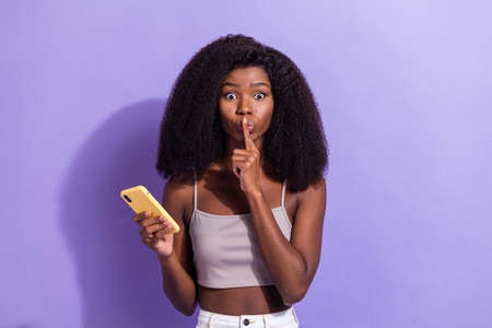 Photo of silent young afro american woman hold finger lips use smartphone isolated on violet color...