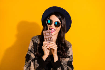 Photo of hungry lady hold bar chocolate open mouth wear specs hat plaid jacket isolated yellow color backgroundの写真素材