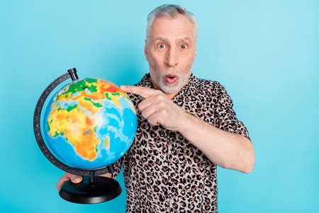 Photo of amazed shocked old man point finger globe tourist destination isolated on blue color...