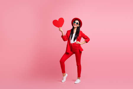 Full length photo portrait of cute girl posing holding big red heart card in one hand isolated on pastel pink colored backgroundの写真素材