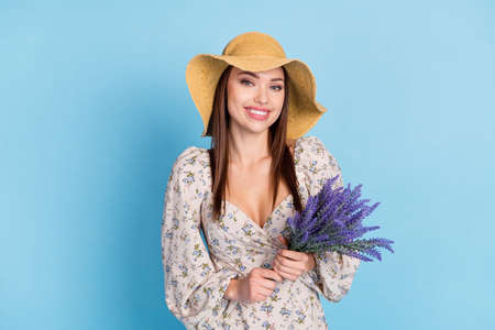 Photo of sweet brown hairdo young lady hold flowers wear hat dress isolated on blue backgroundの写真素材