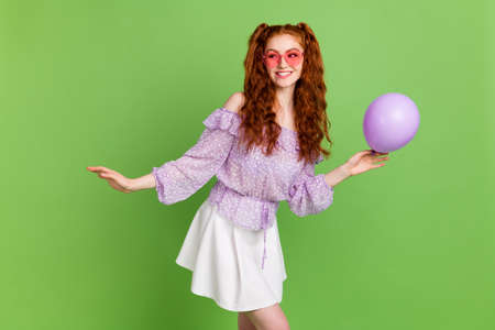 Photo of joyful orange hairstyle young lady hold balloon look empty space wear violet blouse skirt spectacles isolated on green color backgroundの写真素材
