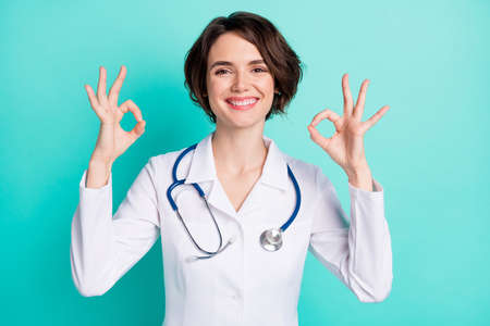 Photo of young woman happy positive smile doctor clinic show okey perfect sign ad isolated over teal color backgroundの写真素材