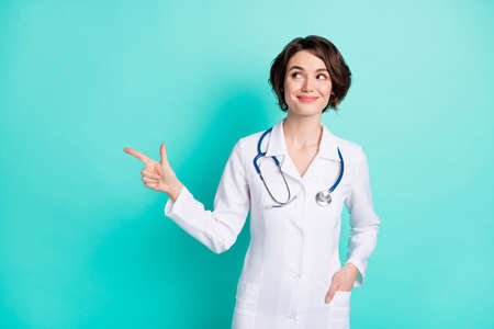 Photo of young woman doctor happy smile look point finger empty space ad promo choose isolated over teal color backgroundの写真素材