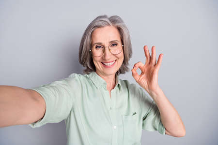 Self-portrait of attractive glad cheerful gray-haired woman showing ok-sign agree ad advert isolated over grey pastel color backgroundの写真素材