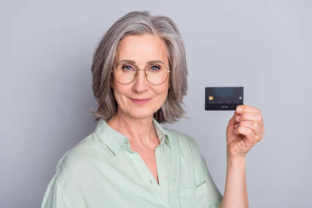 Photo of mature smiling beautiful woman in glasses showing credit card savings salary isolated on grey color backgroundの写真素材