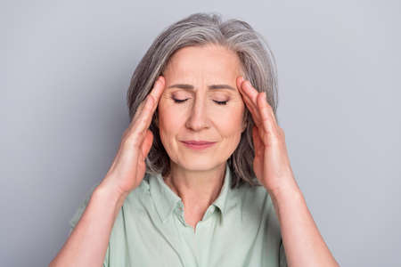 Portrait of attractive depressed gray-haired woman having headache touching temples isolated over grey pastel color backgroundの写真素材