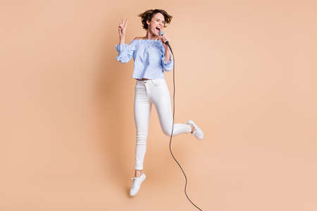 Full length photo of young crazy girl happy sing microphone jump up show cool peace v-sign isolated over beige color backgroundの写真素材