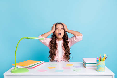 Photo of horrified girl hands on head open mouth look up empty space desk book copybook isolated on blue color backgroundの写真素材