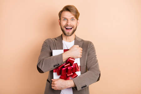 Photo of impressed funny young guy wear jacket smiling hugging gift box open mouth isolated beige color backgroundの写真素材