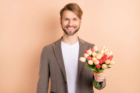 Photo of pretty cute young guy wear jacket smiling holding big tulips bouquet isolated beige color backgroundの写真素材
