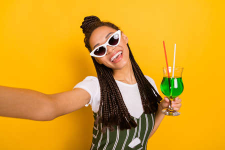 Photo of positive young dark skin woman make selfie camera hold cocktails isolated on yellow color backgroundの写真素材