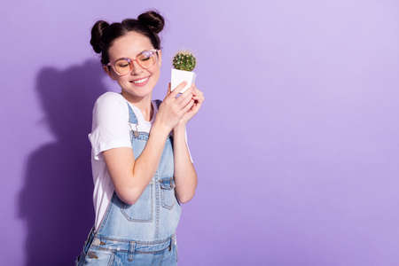 Photo of impressed dreamy young woman dressed denim overall holding small cactus empty space isolated purple color backgroundの写真素材