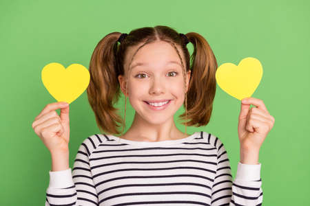 Photo of cute adorable school girl wear white pullover smiling holding two paper hearts isolated green color backgroundの写真素材
