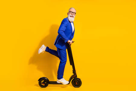 Full body profile photo of funny grey beard aged man ride scooter wear spectacles blue suit isolated on yellow backgroundの写真素材