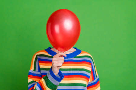 Photo of young woman hold hand cover face balloon hide wear colorful rainbow sweater isolated on green color backgroundの写真素材