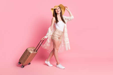 Photo of sweet charming woman dressed white cardigan headwear smiling holding suitcase walking isolated pink color backgroundの写真素材