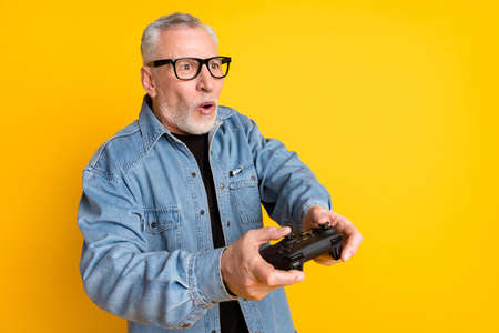 Photo of impressed feel young grandpa play video game wear eyewear jeans jacket isolated on yellow...