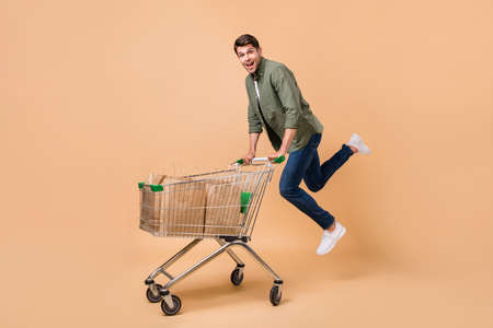 Full body photo of attractive young positive man jump hold trolley bags shop isolated on beige color...
