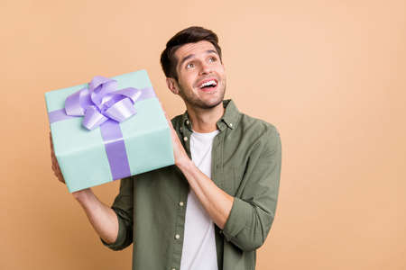 Photo of amazed dreamy young happy man look empty space hold gift box smile isolated on beige color backgroundの写真素材