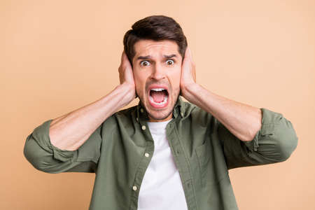 Photo of young amazed shocked afraid young man hold hands cover ears isolated on pastel beige color backgroundの写真素材