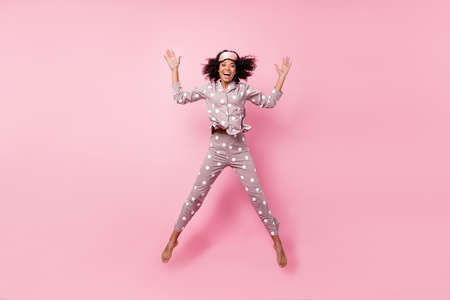 Photo of adorable funny curly dark skin woman dotted nightwear rising arms hands jumping high isolated pink color backgroundの写真素材