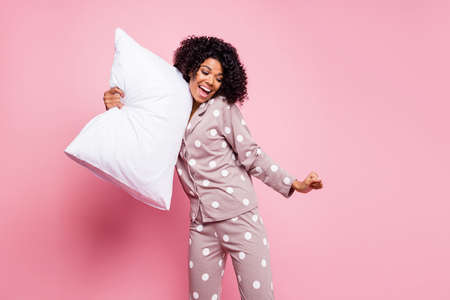 Photo of pretty charming curly dark skin woman dotted nightwear holding white pillow dancing isolated pink color backgroundの写真素材