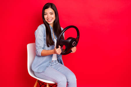 Portrait of pretty cheerful girl sitting in chair holding in hands steering wheel study course isolated over bright red color backgroundの写真素材